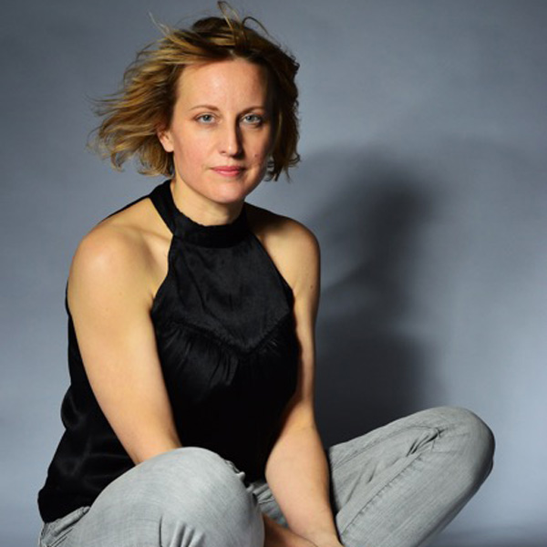 A person with short, light brown hair wearing a sleeveless black top and light gray pants, sitting against a gray background and looking calmly at the camera.