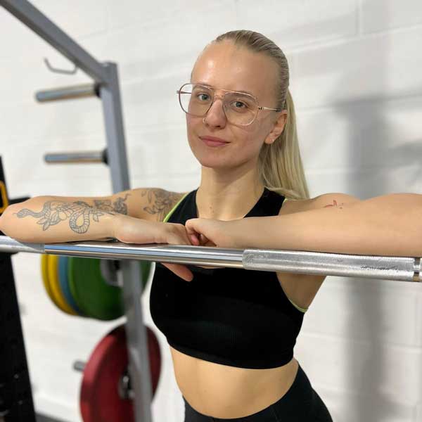 A person with long blonde hair tied back, wearing glasses and a black athletic top, standing in a gym. They are resting their arms on a barbell in a squat rack, with colorful weight plates visible in the background. The setting has a clean, bright atmosphere with a white wall behind them.