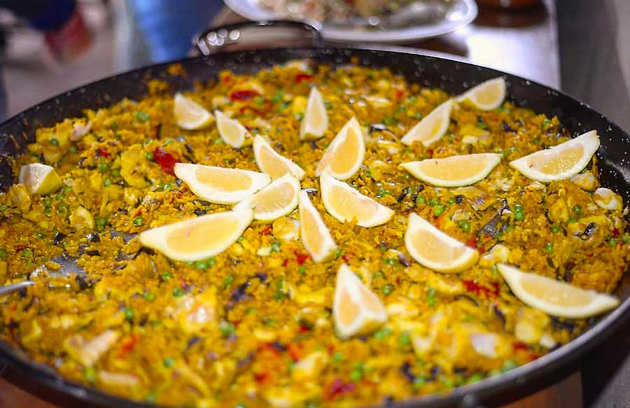 A large pan of vibrant yellow paella garnished with lemon wedges. The dish includes rice mixed with vegetables such as peas and red peppers, along with pieces of seafood or meat, creating a colorful and appetizing presentation.